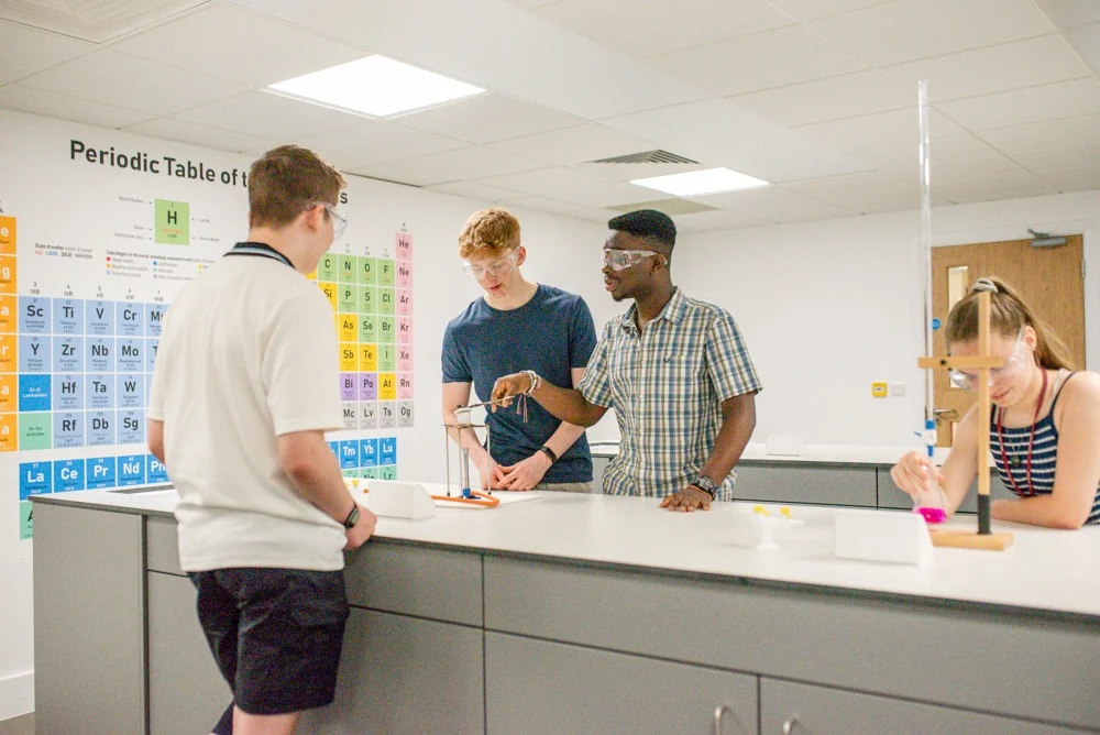 Students conducting a chemistry experiment at a laboratory bench with a periodic table displayed on the classroom wall behind them.